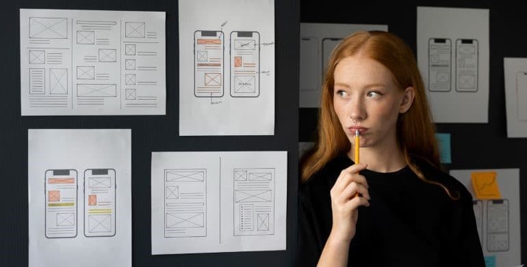 Red-haired female mobile app developer holding a pencil and considering how to reduce expenses.