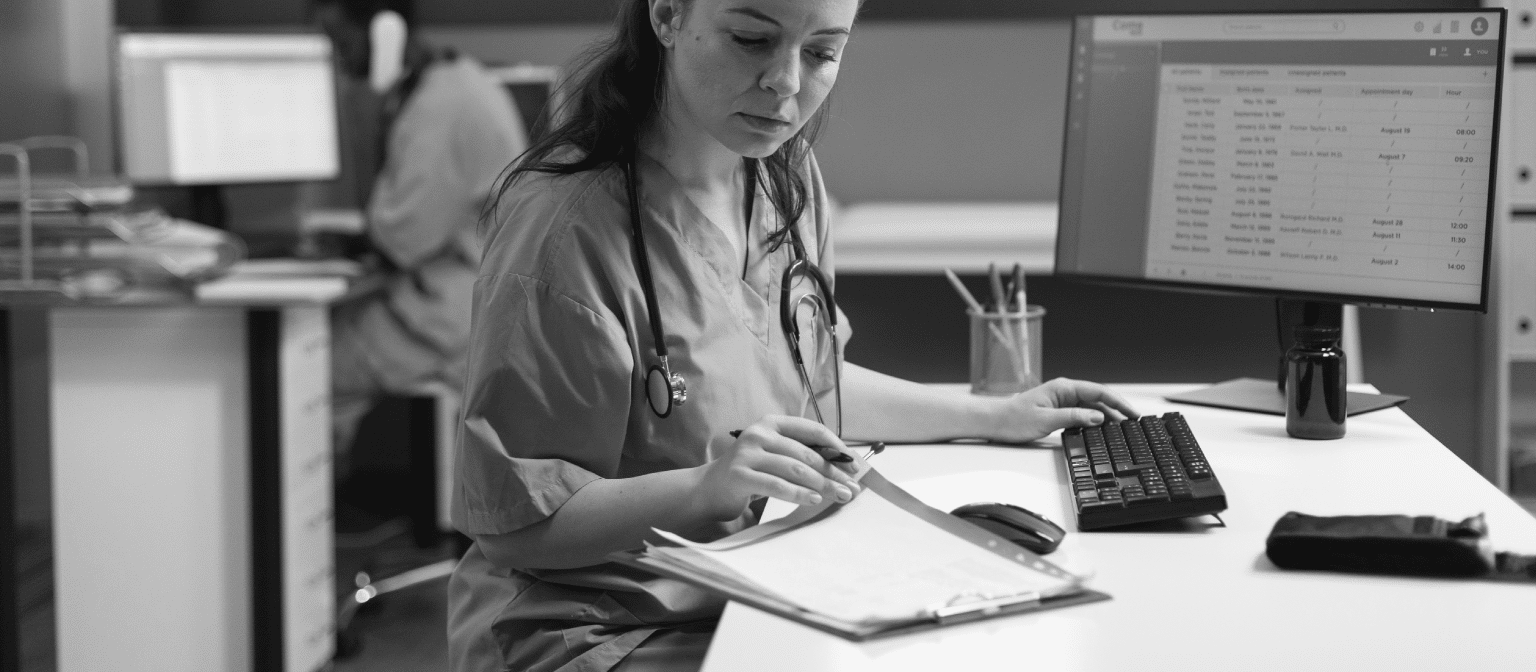 Image of a nurse entering a report in the system while looking at a chart on the table - Healthcare Cyber Security
